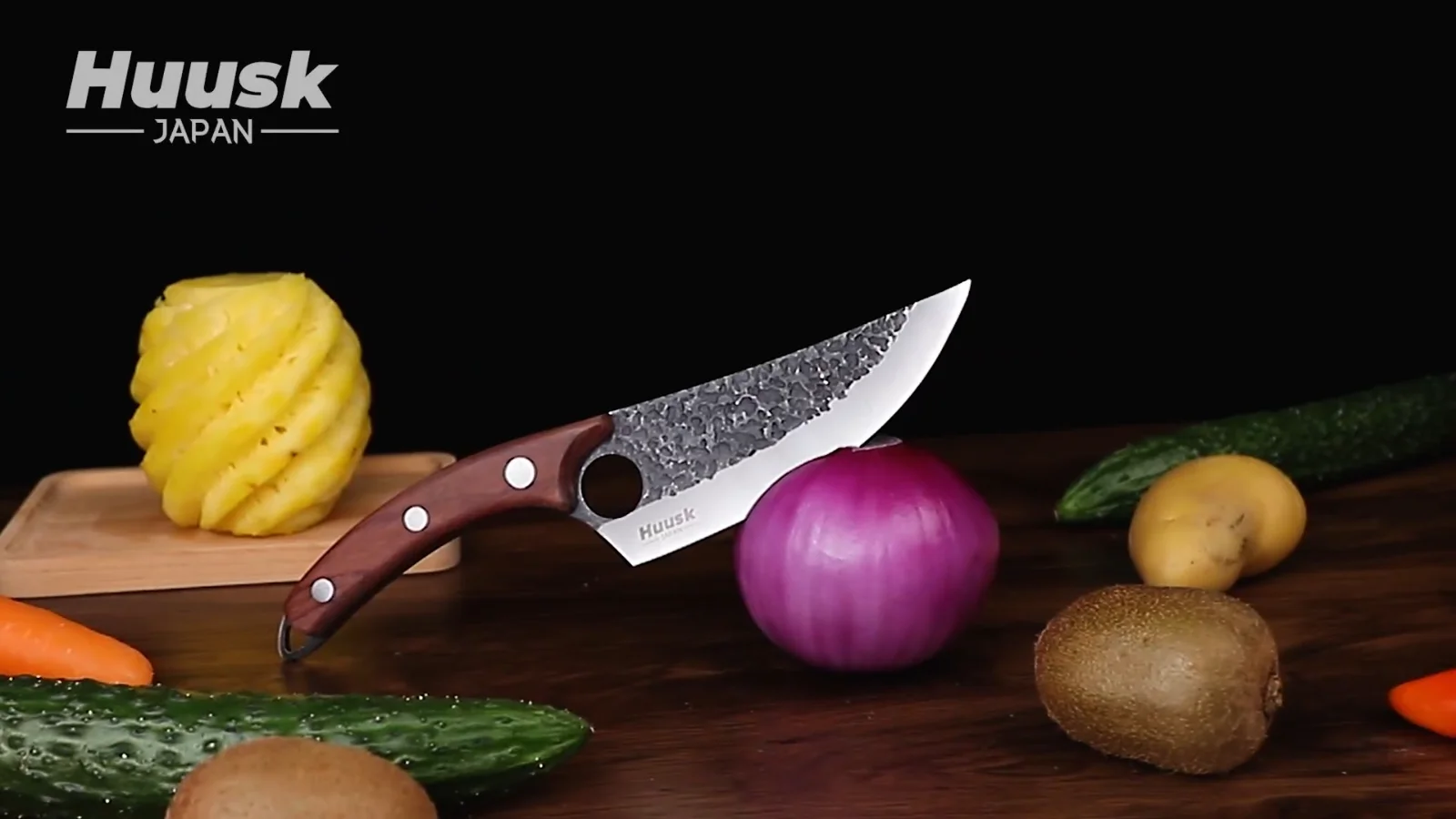 A Huusk hand forged knife lying on a wooden board beside a red onion, carrots and a cucumber